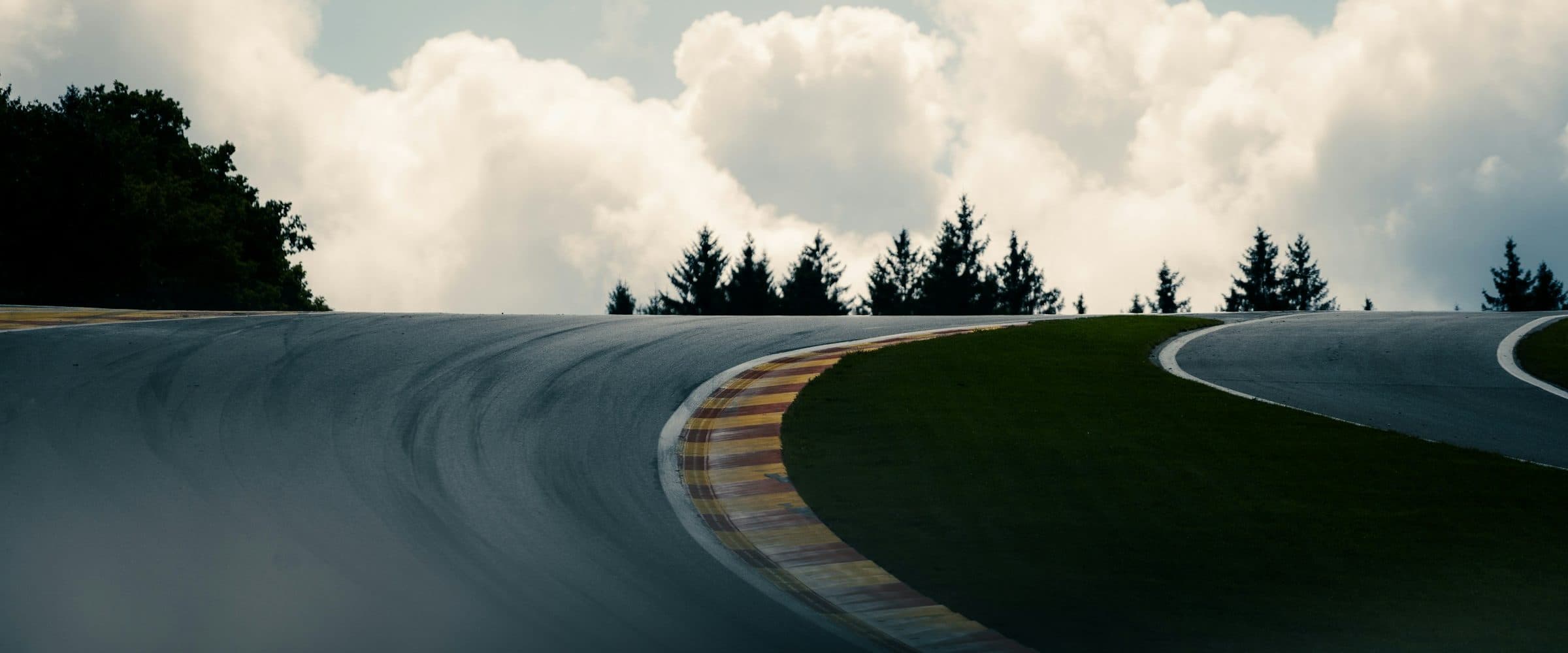 A circuit sweeping downhill past a red and yellow kerb, trees and clouds behind